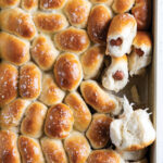 Tray of freshly baked mini sausage rolls in a baking pan, glossy tops with flaky sea salt flakes, some buns showing a sausage filling inside when broken apart