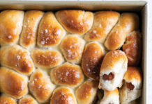 Sausage-Stuffed Dinner Rolls Tray of freshly baked mini sausage rolls in a baking pan, glossy tops with flaky sea salt flakes, some buns showing a sausage filling inside when broken apart