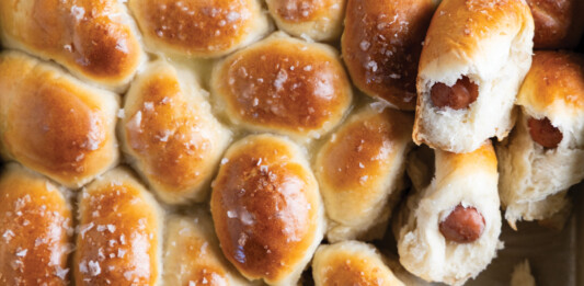 Sausage-Stuffed Dinner Rolls Tray of freshly baked mini sausage rolls in a baking pan, glossy tops with flaky sea salt flakes, some buns showing a sausage filling inside when broken apart