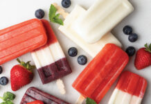 Ice Pops Assorted layered berry popsicles on a white surface, with blueberries, strawberries, and mint garnish nearby.