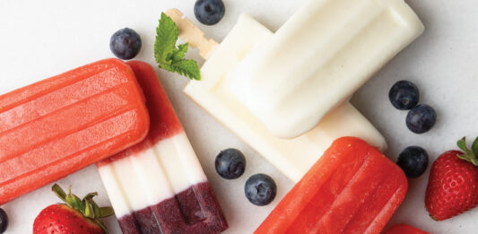 Ice Pops Assorted layered berry popsicles on a white surface, with blueberries, strawberries, and mint garnish nearby.