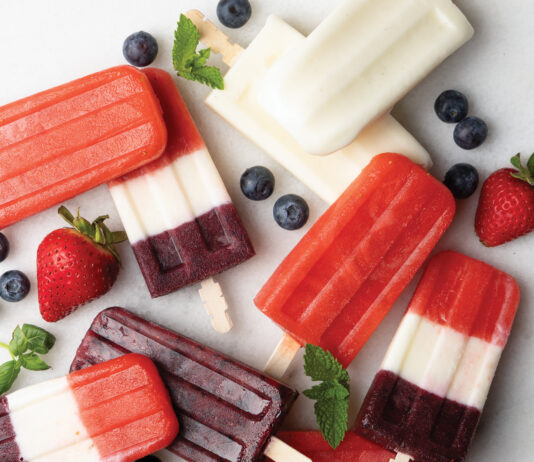 Ice Pops Assorted layered berry popsicles on a white surface, with blueberries, strawberries, and mint garnish nearby.