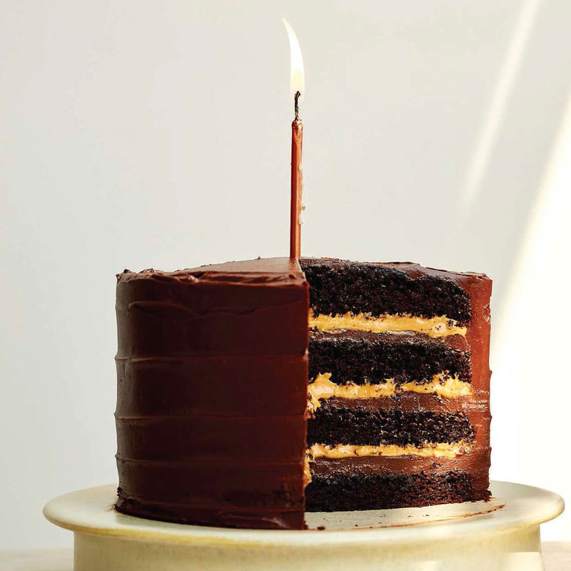 Birthday chocolate cake with a single lit candle on top; slice reveals four layers of chocolate cake with caramel-yellow filling inside.