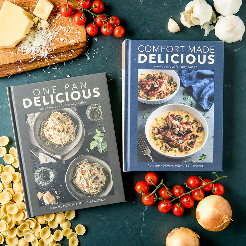 800x800Marketing-8_25JB180 - Bake from Scratch Two cookbooks, 'One Pan Delicious' and 'Comfort Made Delicious,' lie on a dark surface beside pasta, cheese, garlic, onions, and cherry tomatoes for a cozy cooking scene.