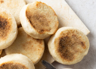 Sourdough English Muffins Stack of toasted English muffins with brown tops on a light board, jam and butter nearby suggests a breakfast spread.