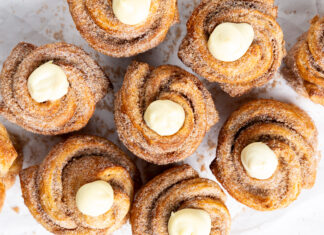 MAPLE CUSTARD MORNING BUNS Cinnamon rolls with a dollop of cream cheese frosting on top, sprinkled with sugar on a parchment surface.