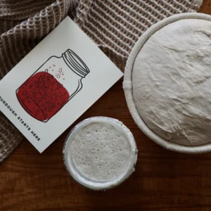Card with a drawn jar of red liquid on a light background, next to an unbaked round pie crust on a wooden table beneath a woven mat