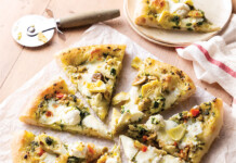 Sourdough Pizza Sliced white pizza topped with artichokes, olives, and herbs on parchment on a wooden table, with a pizza cutter nearby.