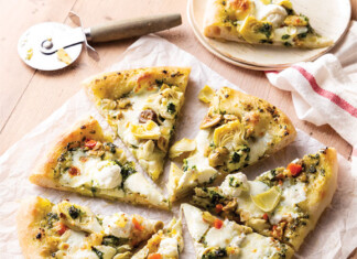 Sourdough Pizza Sliced white pizza topped with artichokes, olives, and herbs on parchment on a wooden table, with a pizza cutter nearby.