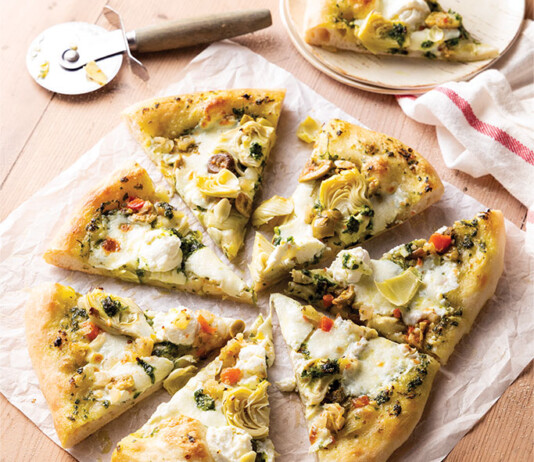 Sourdough Pizza Sliced white pizza topped with artichokes, olives, and herbs on parchment on a wooden table, with a pizza cutter nearby.