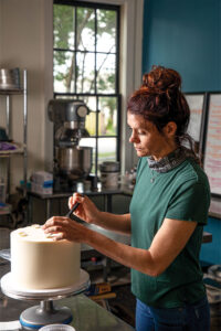 Woman piping icing on a white frosted cake on a rotating stand in a bakery kitchen, with a mixer and shelves in the background.