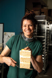 Smiling woman in a green shirt holds a three‑layer cake with white frosting in a bakery.