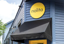 Daily Proof: Nolita Bakery Smiling woman leaning against a bright yellow door outside a blue storefront with a black awning and a Nolita sign.