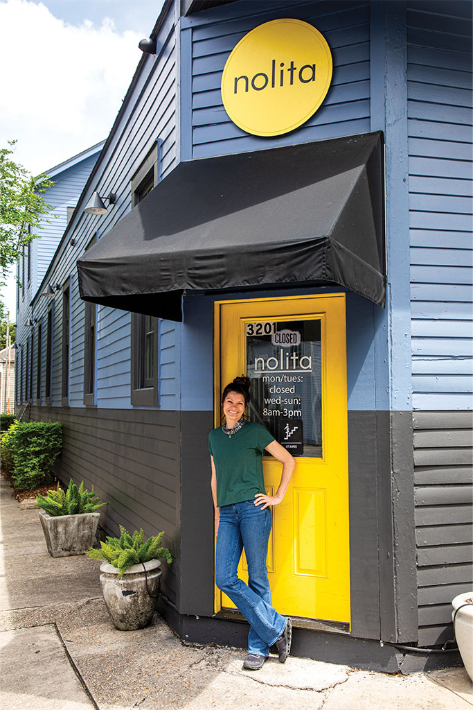 Smiling woman leaning against a bright yellow door outside a blue storefront with a black awning and a Nolita sign.