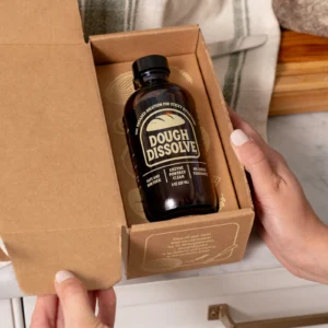 Bottle labeled 'Dough Dissolve' in a cardboard box, held by a person’s hands, showing its dark brown container and black cap.
