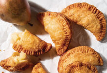 Honey-Vanilla Pear Hand Pies Assorted baked fruit-filled hand pies (empanadas) on parchment with a pear, a halved apple, and dipping sauce nearby.