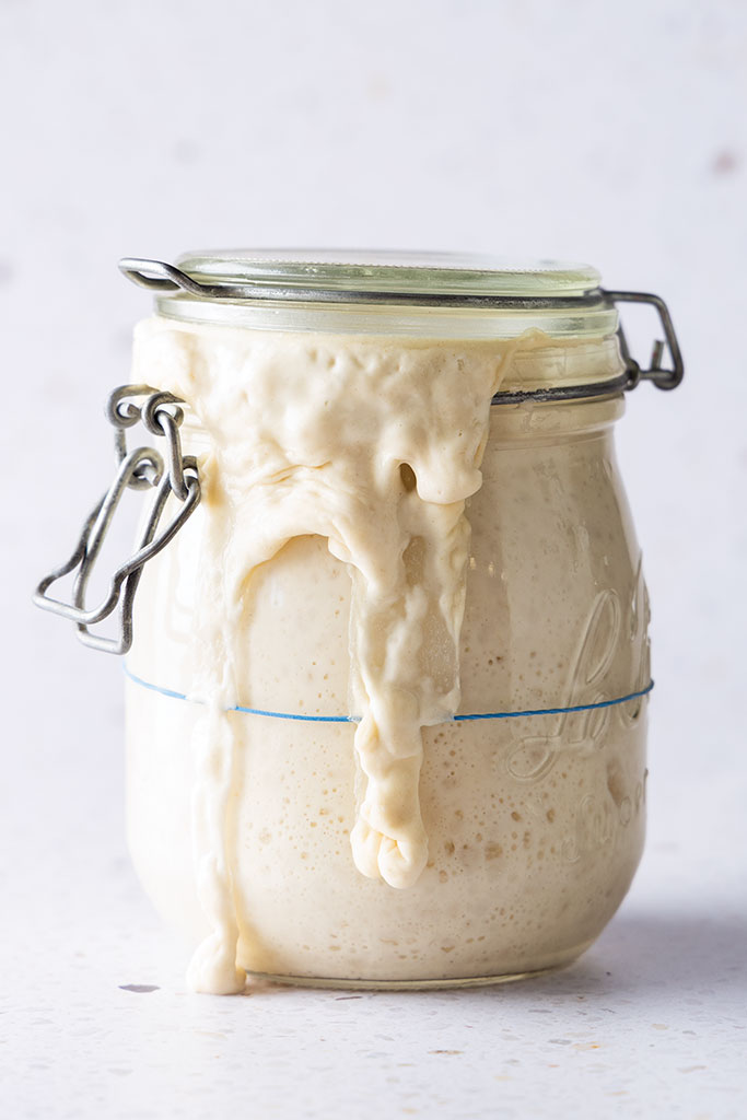 Mason jar of sourdough starter overflow­ing with foamy, pale batter on the rim and down the side.