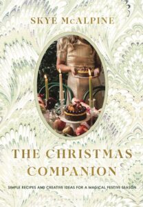 Book cover for The Christmas Companion by Skye McAlpine; an oval photo shows a woman in a gold dress holding a cake, set on a marbled pastel background.