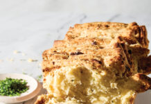 Pull-Apart Chive-Feta Biscuit Loaf Sliced rustic bread stacked on a white plate with a crumbly golden crust and herbs nearby.