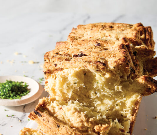 Pull-Apart Chive-Feta Biscuit Loaf Sliced rustic bread stacked on a white plate with a crumbly golden crust and herbs nearby.