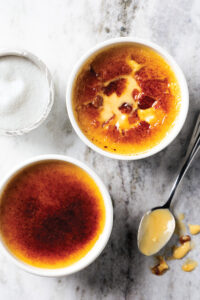 Two ramekins of crème brûlée with caramelized tops on a marble countertop, sugar bowl in the background.