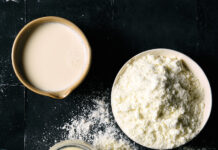 From the Pantry: The Three Milks Top-down view of a mug of milk and a bowl of flour on a black tiled surface, with flour specks scattered nearby.