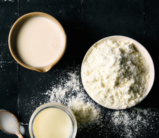 From the Pantry: The Three Milks Top-down view of a mug of milk and a bowl of flour on a black tiled surface, with flour specks scattered nearby.