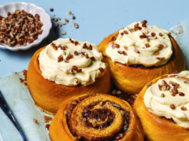 Jumbo Pumpkin-Pecan Sweet Rolls Golden cinnamon rolls topped with cream cheese frosting and chopped pecans on a blue surface.