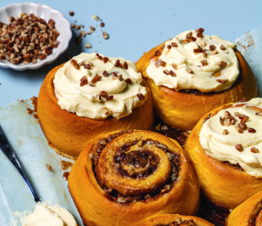 Jumbo Pumpkin-Pecan Sweet Rolls Golden cinnamon rolls topped with cream cheese frosting and chopped pecans on a blue surface.